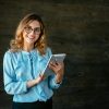 Happy beautiful young business woman standing with tablet, widely smiling. Dressed in elegant blouse, in eyeglasses.